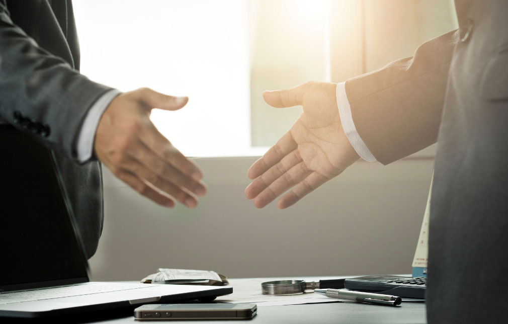  Two professionals in suits reaching out for a handshake over a desk.
