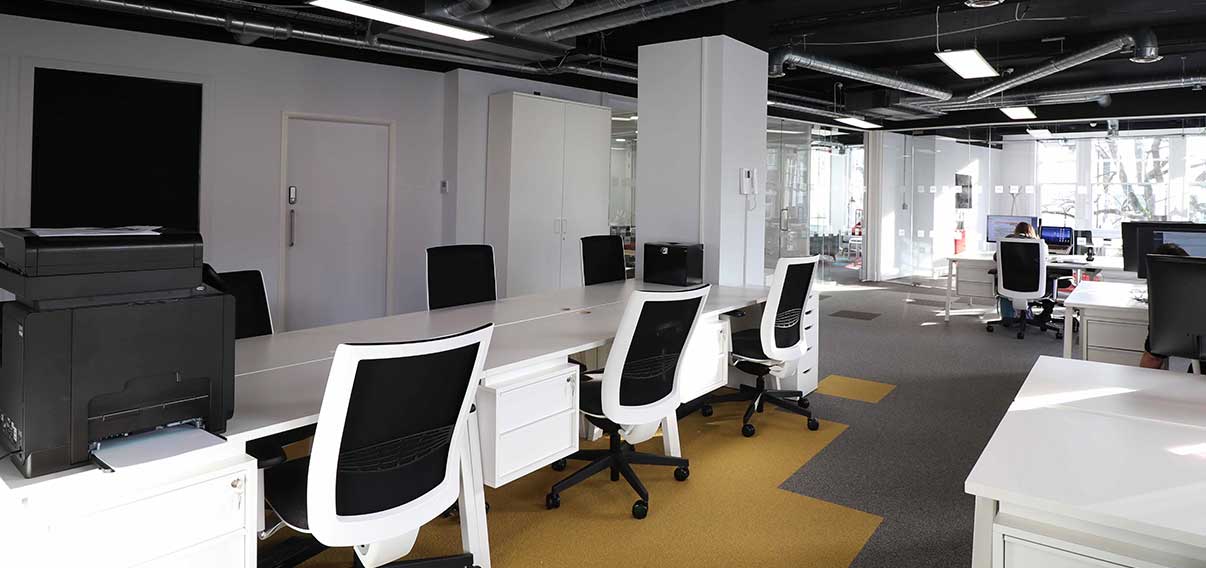 Contemporary office featuring white desks, black and white chairs, a multifunction printer, glass partitions, and a contrast of gray and yellow floors, illuminated by natural light with exposed black ceiling ducts.