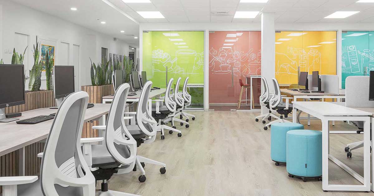 Modern office space with rows of workstations, desktop computers, white chairs, wooden partitions, and plants. Glass walls with green and orange panels feature white line drawings, complementing the light-colored floors and walls.